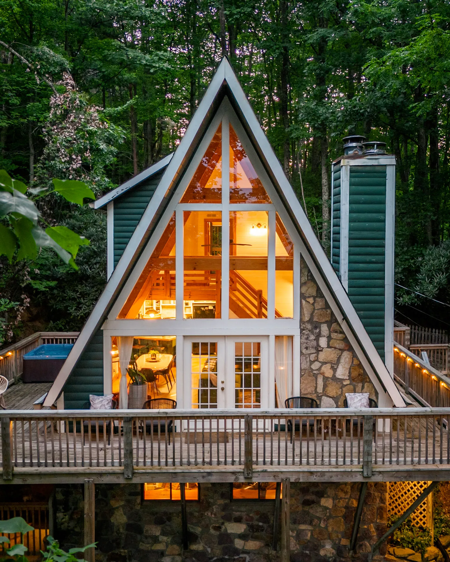 Little Green Cabin, a green A-frame with stone chimney, lit from within at dusk